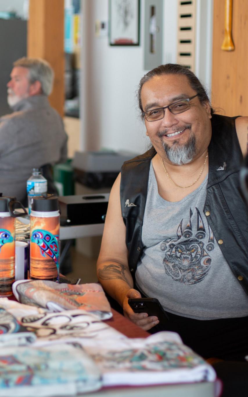 Indigenous man at table covered in craftwork of various kinds. 
